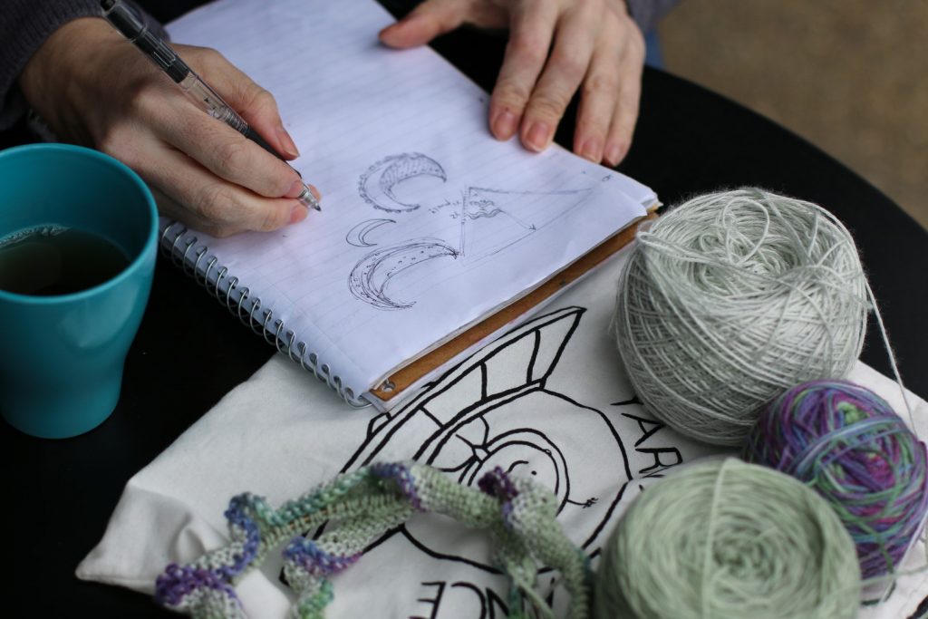 A close up of hands making design notes in a notebook, surrounded by balls of green yarn and some knitting, to indicate that SpaceCadet is looking for test knitters and crocheters